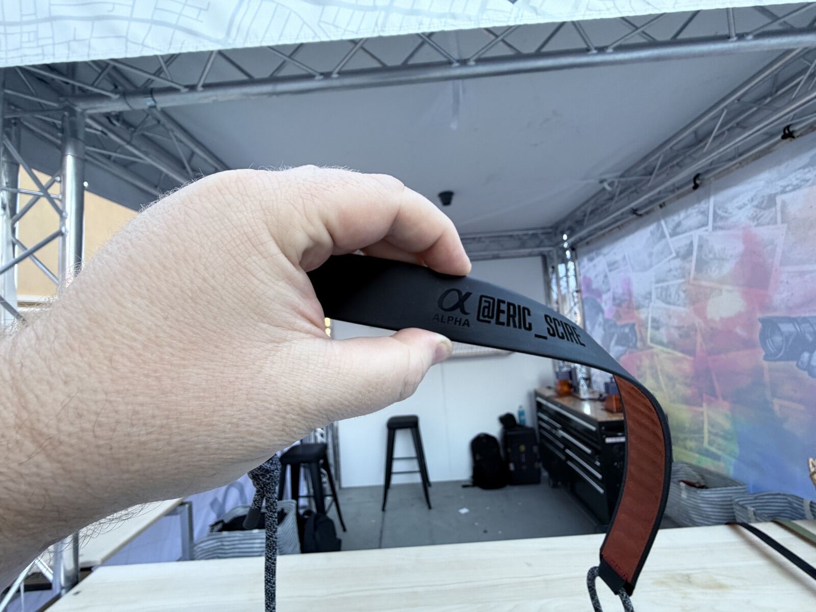 Hand holding a black camera strap freshly laser engraved with a guest's social media handle at an outdoor brand festival booth