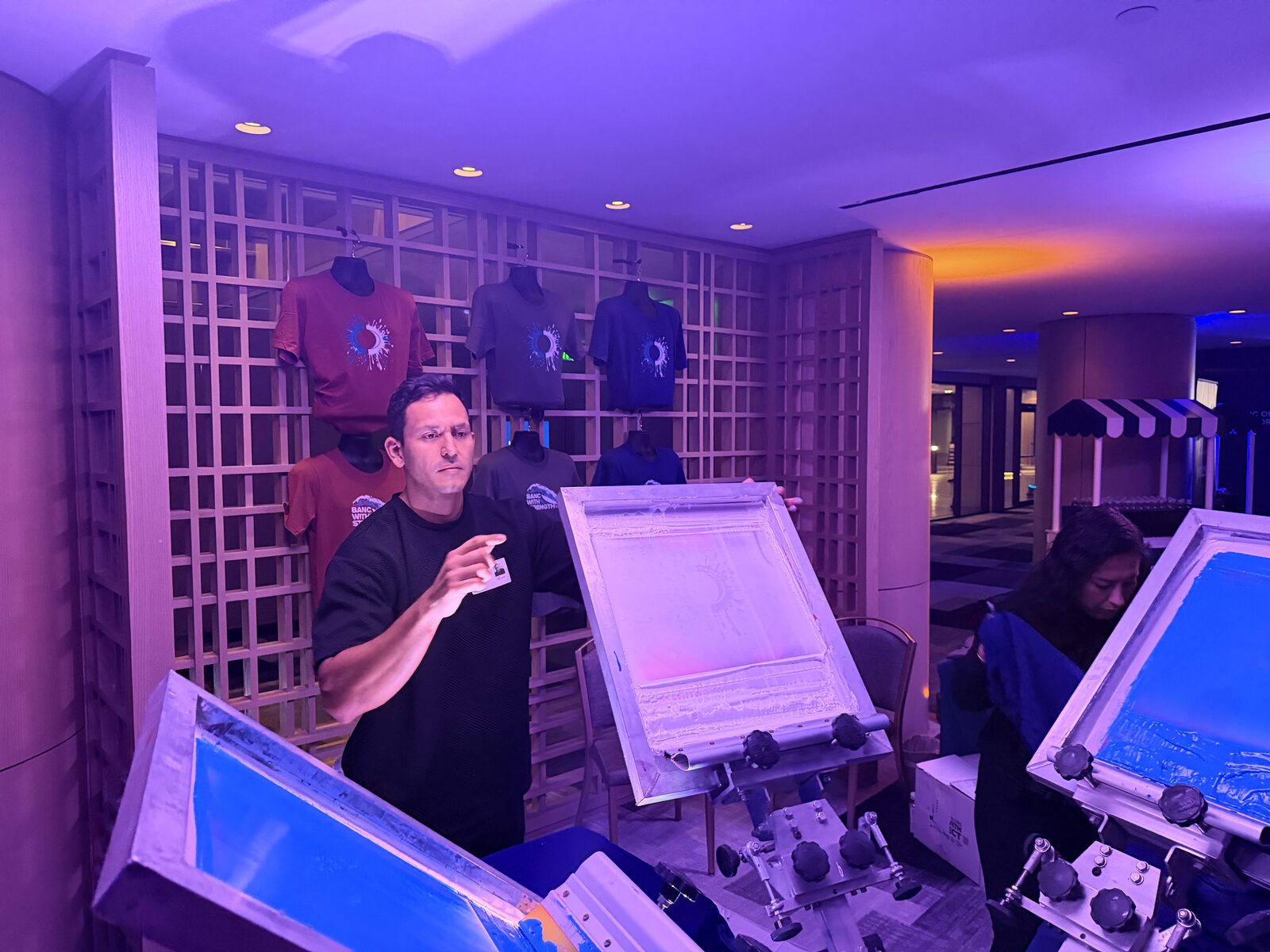 Merch Troop live printing stations running under evening lighting at a hotel event with finished shirts displayed behind the crew